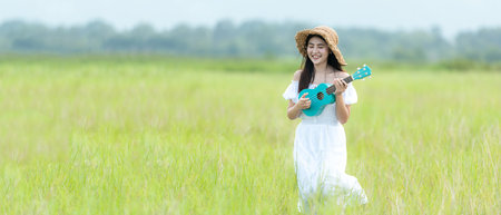 Lifestyle asian women white dress holding a guitar on a cloudy sunrise sky in the meadow flower, relax and happy day on summertime, outdoors background field copy space for bannerの写真素材