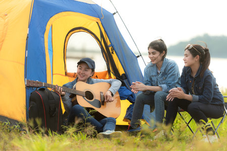 Camping outdoor. Group  friends camping leisure and destination travel. Family sitting around camp fire and playing guitar and roasted sausages, tourism relax and travel near riverの写真素材