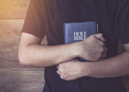 a young man in black t-shirt holding holy bible in his arms over wooden backgroundの写真素材