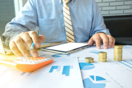 businessman using calculator with stacked coins arranged at office desk and many document data graph...
