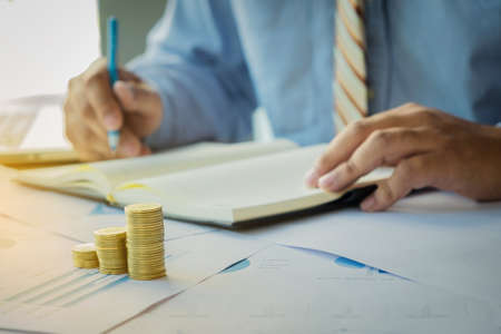 businessman writting note paper with stacked coins arranged at office desk and many document data graph in morning light, business concept.の写真素材
