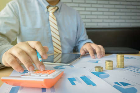 businessman using calculator with stacked coins arranged at office desk and many document data graph in morning light, business concept.の写真素材