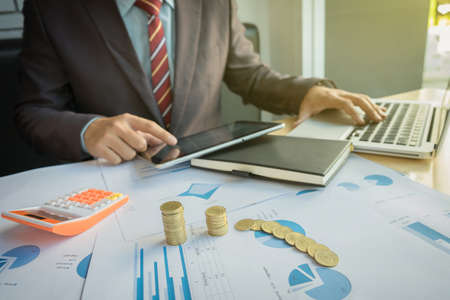 businessman using tablet and calculator with stacked coins arranged at office desk and many document data graph in morning light, business concept.の写真素材
