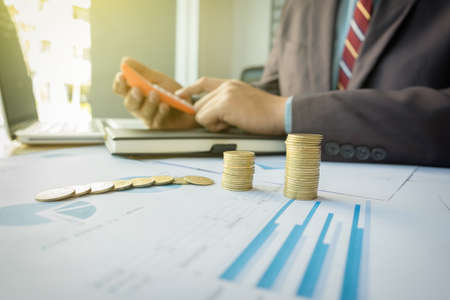 businessman using calculator with stacked coins arranged at office desk and many document data graph in morning light, business concept.の写真素材