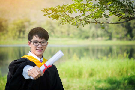 Happy graduate. Happy Asian man in graduation gowns holdin on nature background.の写真素材