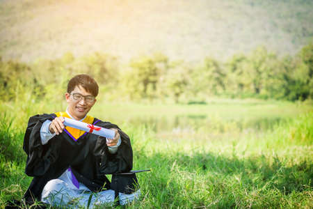 Graduation: Student sit and smile holding certificate, graduation hat with Diploma With background in the nature.の写真素材