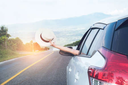 Relaxed happy traveler, Young beatiful asian gilr holding white Hat weave and Reach out of the car at sunset and beautiful view with mountain road background.の写真素材