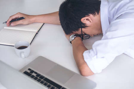 Tired businessman sleeping while working with laptop and writing book at work in modern office, people dozing on workplace.の写真素材