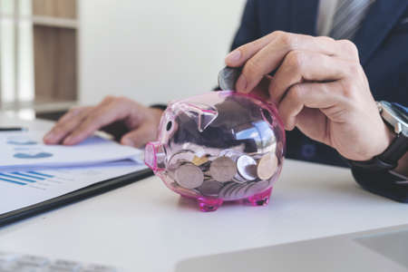 Businessman putting coins into piggy bank and using calculator to analysis business investment with many document graph data, financial concept.の写真素材