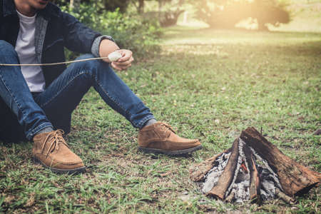 Young Man hiking cooking a marshmallow candies on the campfire in forest, hike and people concept -...