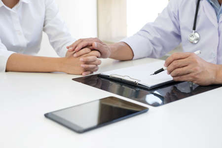 Friendly Professional medical doctor in white uniform coat holding patient hand sitting at the desk for encouragement, cheering and support while listen medical results.の写真素材