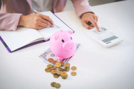 Business woman putting coins into piggy bank, writing to report and using calculator to analysis business investment strategy with income of money coin and dollar, financial concept.の写真素材