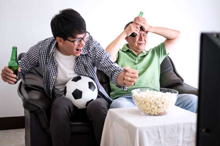 Young Asian Man and father watching soccer match on tv and cheering football team, celebrating with beer and popcorn at home, sports and entertainment concept.の写真素材