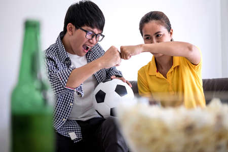 Young Asian couple love watching soccer match on tv and cheering football team, celebrating with beer and popcorn at home, sports and entertainment concept.の写真素材
