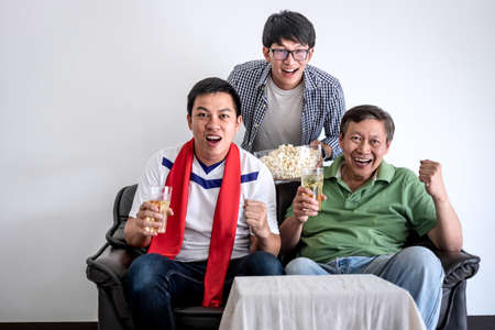 Young Happy Asian Men family or football fans watching soccer match on tv and cheering football team, celebrating with drink beer and eat popcorn at home, sports and entertainment concept.の写真素材