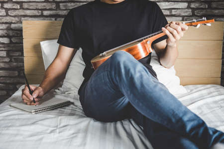 Young happiness man on bedroom in enjoying playing the music and compose a song with guitar or ukulele in holiday, relaxation and recreation concept.の写真素材