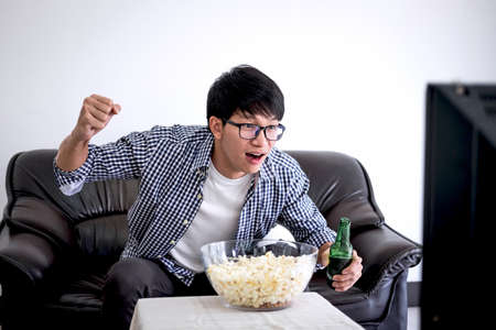 Young Asian Man fanclub watching soccer match on tv and cheering football team, celebrating with beer and popcorn at home, sports and entertainment concept.の写真素材