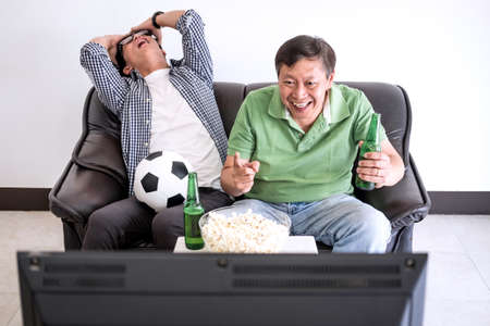 Young Asian Man and father watching soccer match on tv and cheering football team, celebrating with beer and popcorn at home, sports and entertainment concept.の写真素材