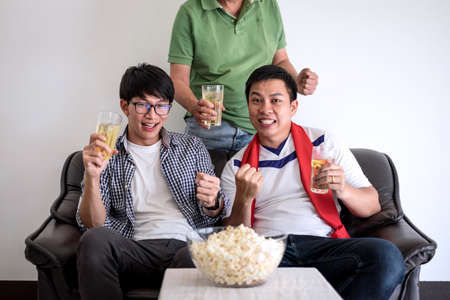 Young Happy Asian Men family or football fans watching soccer match on tv and cheering football team, celebrating with drink beer and eat popcorn at home, sports and entertainment concept.の写真素材