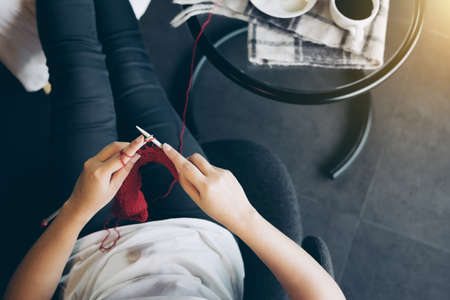 Close up shot of young woman hands knitting a red scarf handicraft in the living room on terrace at...