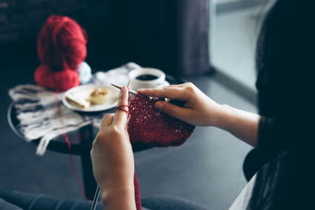 Close up shot of young woman hands knitting a red scarf handicraft in the living room on terrace at...