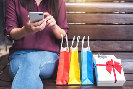 Consumerism, shopping, lifestyle concept, Young woman sitting near shopping bags and gift box while playing smartphone enjoying in shopping.の写真素材