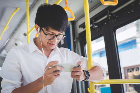 Young Asian man traveler standing on a bus using smartphone watch video or playing game while smile of happy, transport, tourism and road trip concept.の写真素材