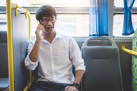 Young Asian man traveler sitting on a bus talking and using smartphone while smile of happy, transport, tourism and road trip concept.の写真素材