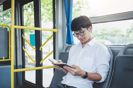 Young Asian man traveler sitting on a bus listening music and reading book while smile of happy,...
