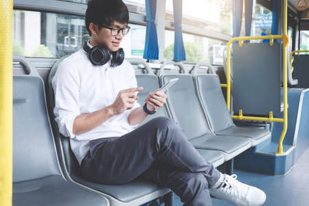 Young Asian man traveler sitting on a bus using smartphone watch video or listening music while smile of happy, transport, tourism and road trip concept.の写真素材