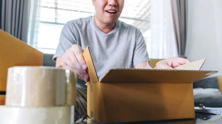 Small business parcel for shipping, Happy man opening online shopping package box with parcel while sitting on sofa at home.の写真素材