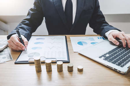 Businessman writing making report with stacked coins arranged at office desk and many document data graph, business concept.の写真素材