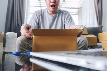 Small business parcel for shipping, Happy man opening online shopping package box with parcel while sitting on sofa at home.の写真素材