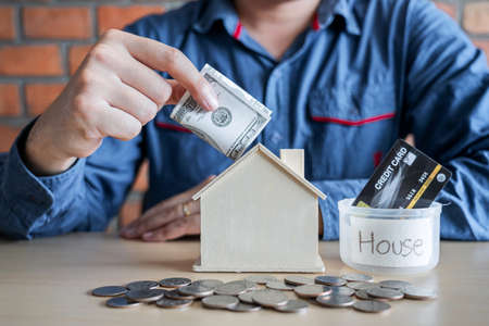 Man putting coins into money box with savings and analysis business investment strategy home loan with income of money coin and dollar for future real estate.の写真素材