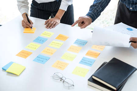 Two creative business people meeting and planning use sticky note on desk to share idea, Analysis data chart and graph with teamwork strategy brainstorming in office.の写真素材