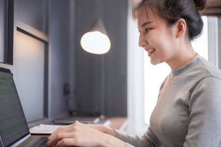 Close up of young asian woman in casual clothes looking on laptop screen and typing financial project about online business on laptop in cozy workplace at homeの写真素材