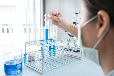 Scientist woman in medical face mask is holding test tube with blue liquid and looking reaction of experimental  vaccine while working with microscope in laboratoryの写真素材