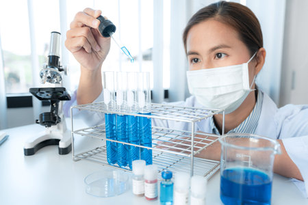 Scientist woman in medical face mask is holding dropper to dropping chemical liquid into test tube and looking reaction of experimental  vaccine while working in laboratoryの写真素材