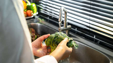 Close up of asian housewife wearing an apron and standing to washing broccoli and vegetables with...