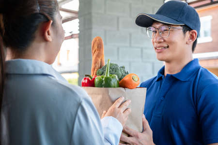 Woman customer standing at the house to accepting a grocery bag of food, fruit, vegetable from asian delivery man in blue uniform that service delivery during coronavirus covid-19 pandemicの写真素材