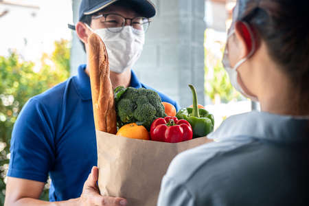 Asian delivery man in blue uniform with medical face mask sending grocery bag of food, fruit, vegetable to woman customer in front of the house that service delivery during covid-19 pandemicの写真素材