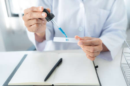 Scientist woman holding dropper to dropping solution sample on the glass plate for microscope and blue liquid in test tube while working to analyzing and developing coronavirus vaccine in laboratoryの写真素材