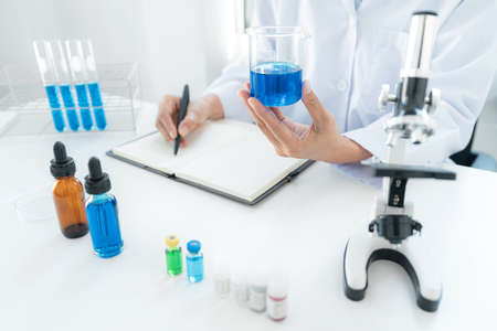Scientist woman holding beaker in his hands and writing down result report on notebook while working...