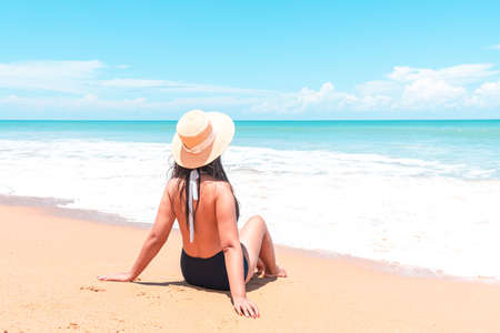 Back view of asian traveler woman in swimsuit and straw hat to sitting in the spray of the sea to looking view the blue horizon on the sea while relaxing in holiday tripの写真素材
