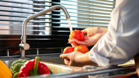 Hands of asian housewife washing tomato and vegetables with water in sink while wearing apron and...