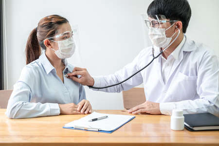 Male doctor in medical mask is using stethoscope to examining heartbeat and symptom of patient while talking about disease symptom and explaining treatment of the illness in clinicの写真素材