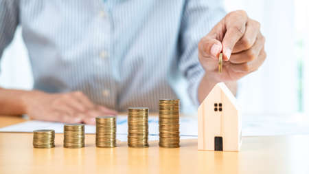 Asian business woman holding coin to putting into house piggy bank with increasing coins stack for...