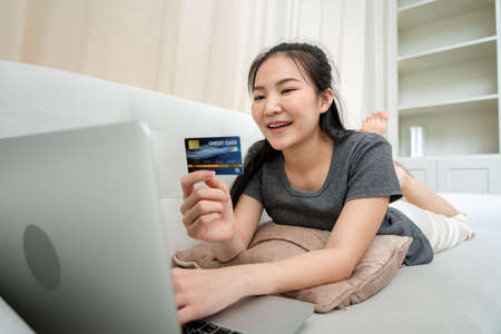 Freelance woman holding credit card for enter information to receive payment and check balance in banking app while lying on couch to receive order from client during online marketing delivery in homeの写真素材