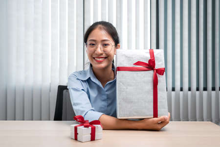 Happy woman is smiling and holding big present box with red ribbon in the hands while preparing gift...