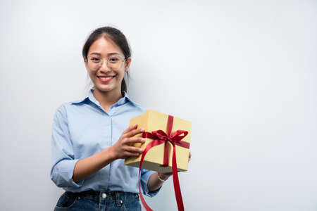 Happy woman is smiling and holding present box with red ribbon in the hands while standing to...